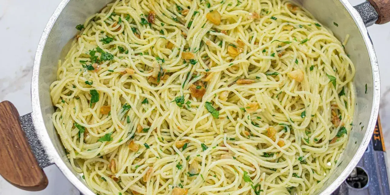 Espaguetis al ajillo recién hechos, con trocitos de ajo dorado y perejil fresco, servidos en una sartén metálica.