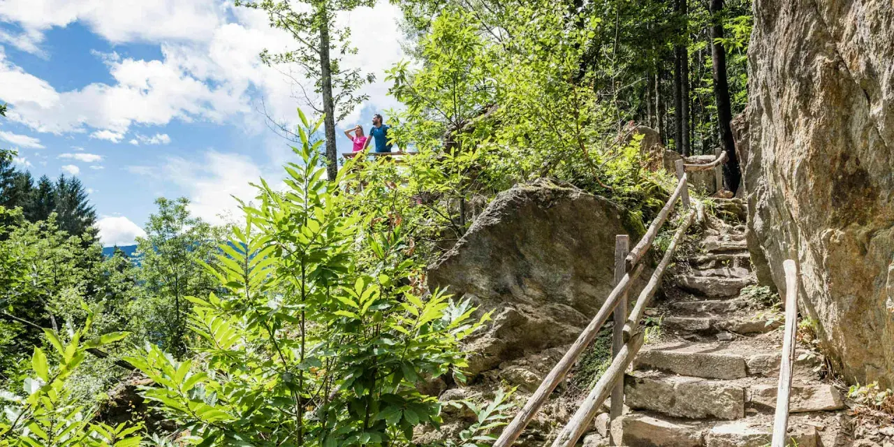 Ein Paar genießt die Aussicht vom Schenner Waalweg. Steintreppen führen durch üppige Vegetation und Felsen.