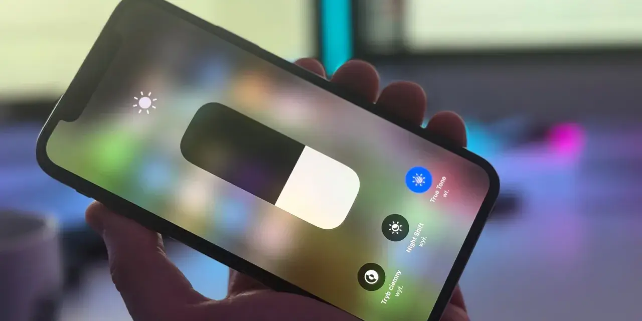 Dłoń trzyma iPhone'a z włączonym panelem sterowania. Widoczny suwak jasności i ikona True Tone.