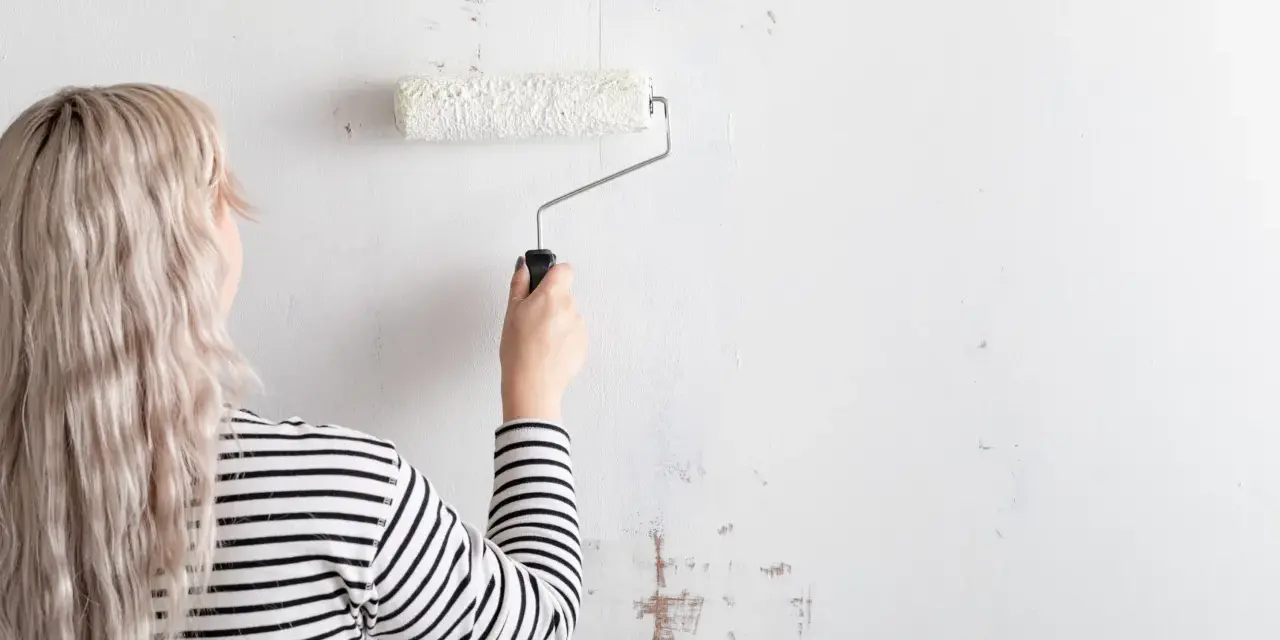 Mujer con cabello rubio y camiseta a rayas está a punto de empapelar pared con rodillo blanco.