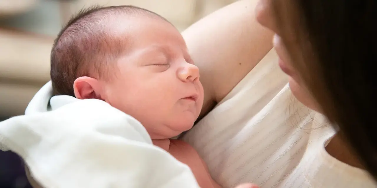 Śpiące niemowlę w ramionach mamy. Warto sprawdzić, ile powinno jeść niemowlę -- tabela, by zapewnić mu najlepszy start.