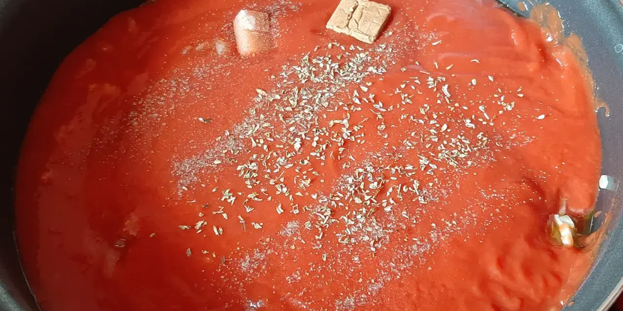 Un coulis de tomates mijote dans une poêle, agrémenté d'herbes et d'un cube de bouillon.