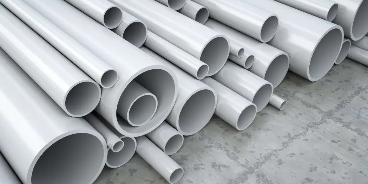 A pile of white pipes, many of which are made of vinyl, a type of plastic, are stacked on a concrete floor.