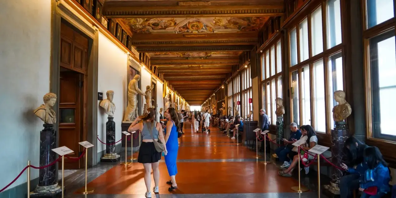 Wnętrze muzeum Uffizi z rzeźbami i malowanym sufitem. Ludzie spacerują i odpoczywają wzdłuż korytarza.