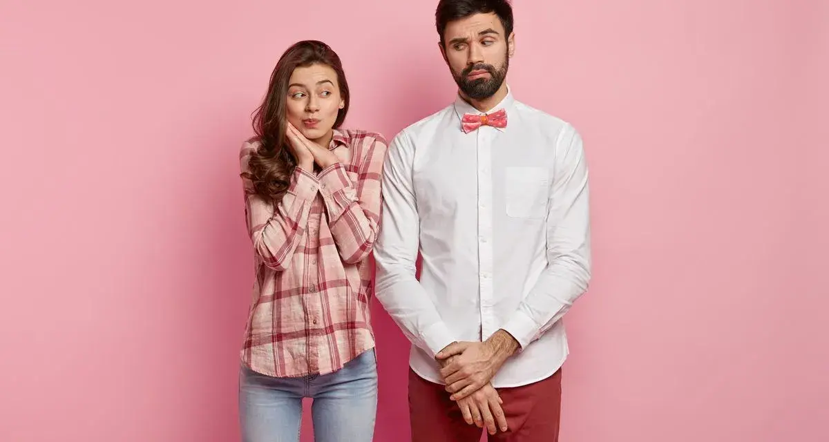Coppia su sfondo rosa. Lei, con camicia a quadri e jeans, sembra timida. Lui, con camicia bianca e papillon, è pensieroso. Come vestirsi al primo appuntamento uomo?