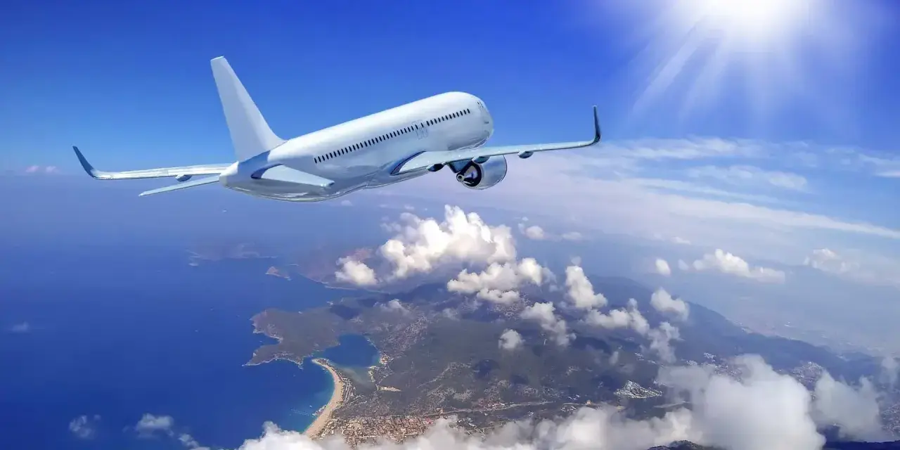 Un aereo bianco solca il cielo azzurro sopra una costa frastagliata. Il sole splende, creando un senso di distacco del vitreo e aereo.
