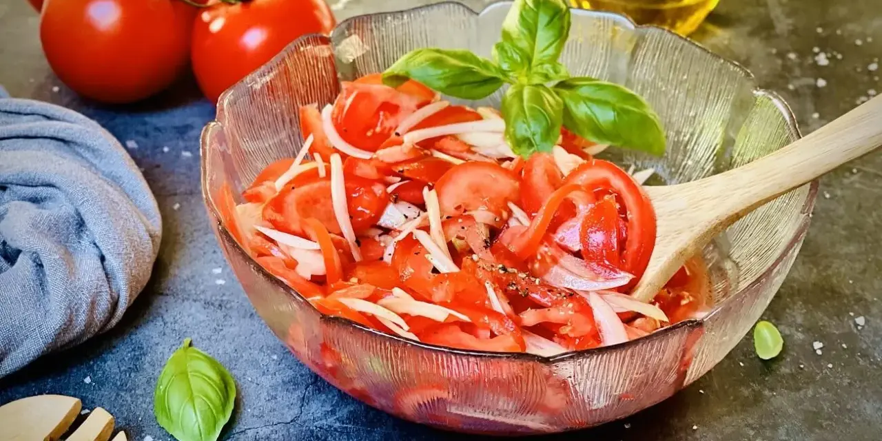 Frischer tomatensalat mit zwiebeln, garniert mit Basilikum. Daneben liegen frische Tomaten und ein Holzlöffel.