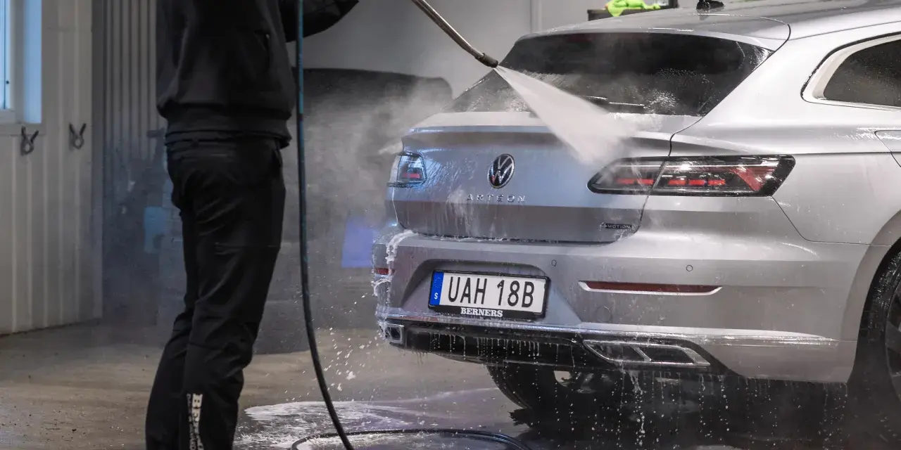 En person tvättar en silverfärgad bil med högtryckstvätt. Frågan "får man tvätta bilen hemma" dyker upp när man ser detta.