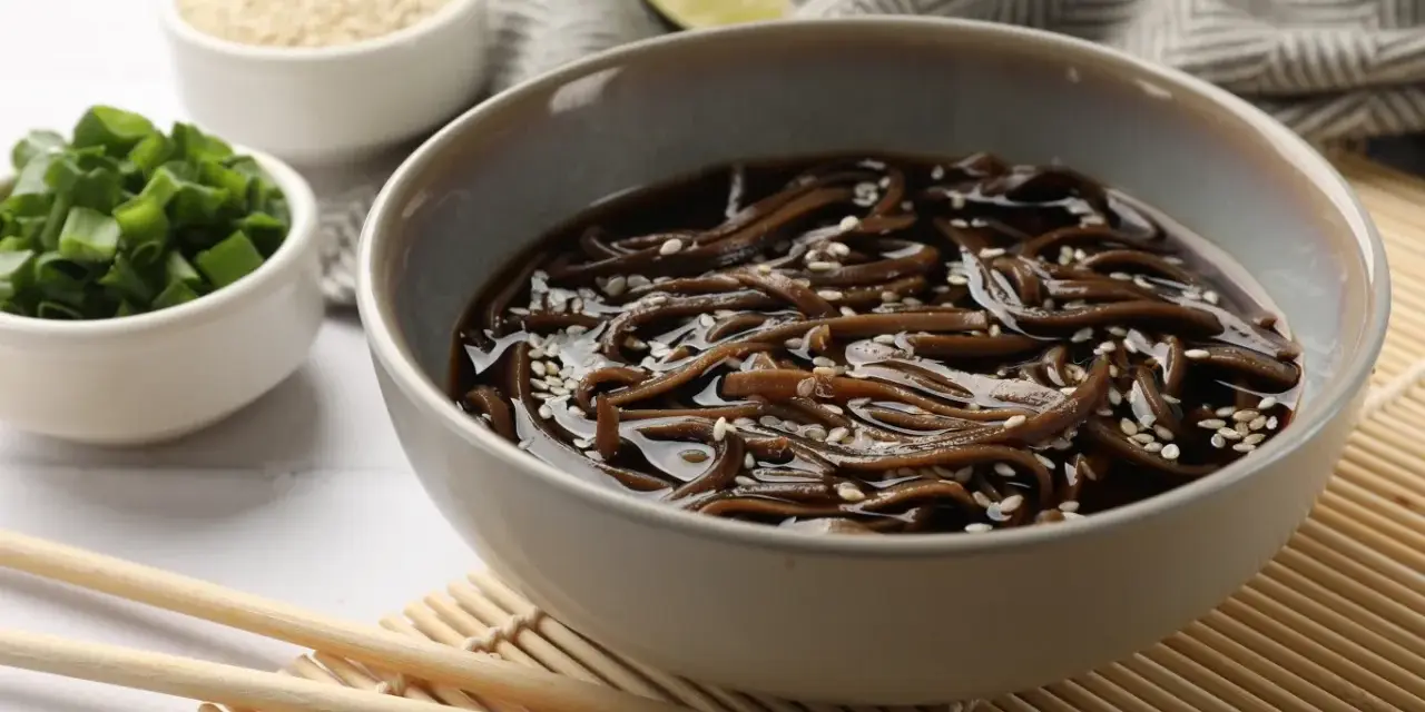 Miseczka z makaronem soba w sosie, posypana sezamem. Obok miseczki z posiekanym szczypiorkiem i sezamem. Zdrowy posiłek, mało makaron soba kcal.
