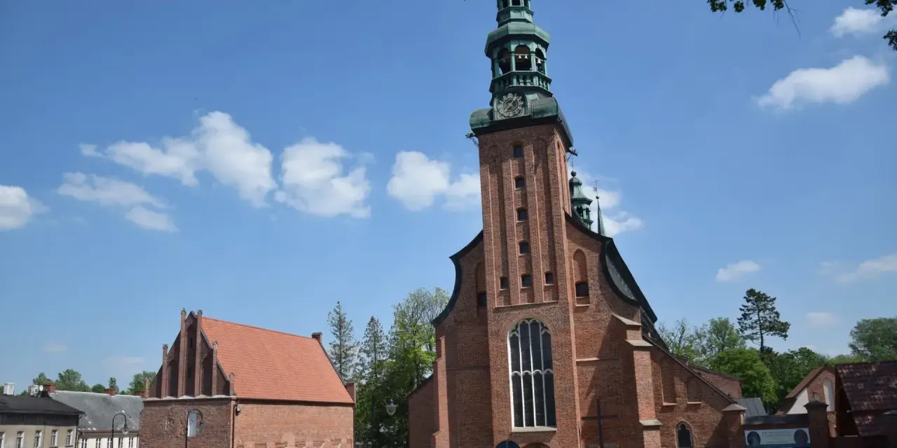 Majestatyczna kolegiata w Kartuzach z cegły, z zieloną wieżą i zegarem, na tle błękitnego nieba z chmurami.
