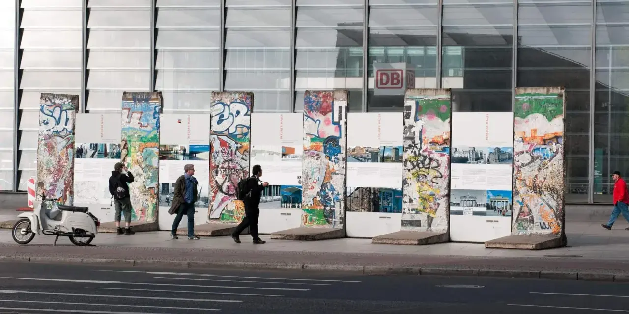 Bunte Reste des Berliner Mauer-Denkmals vor dem Bahnhof Potsdamer Platz. Menschen spazieren vorbei, ein Roller parkt daneben.