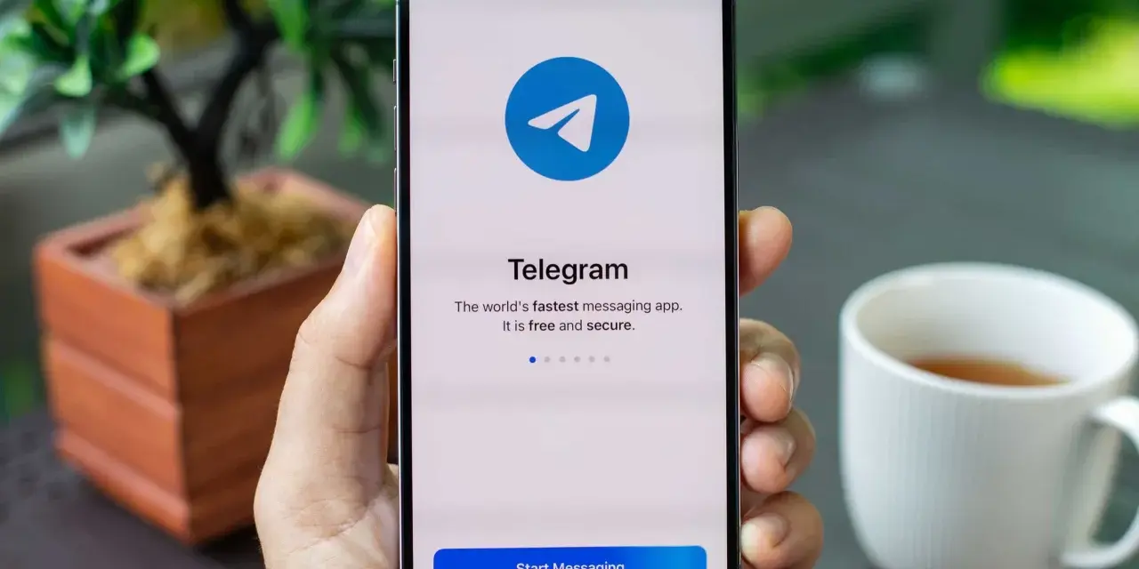 Dłoń trzyma smartfon z otwartą aplikacją Telegram. Na ekranie widnieje logo i napis "Telegram. The world's fastest messaging app. It is free and secure.