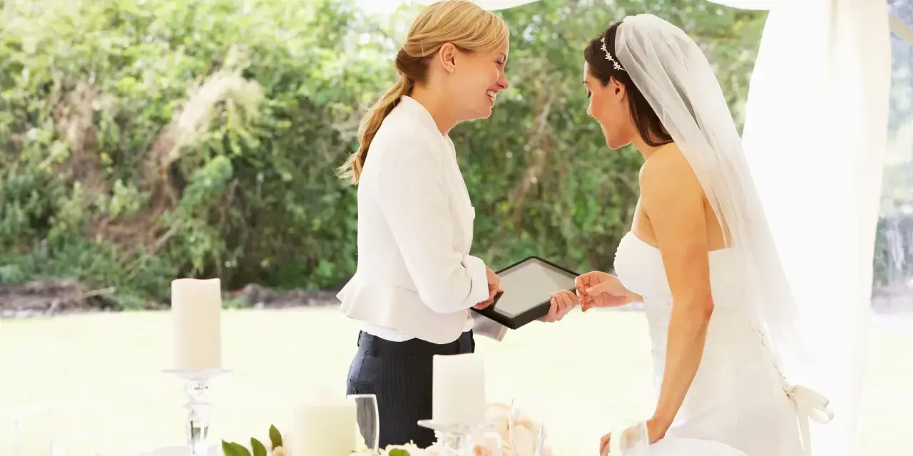 Panna młoda z welonem i wedding plannerka dyskutują przy stole. Czy warto wynająć wedding plannera, czy poradzić sobie samemu?