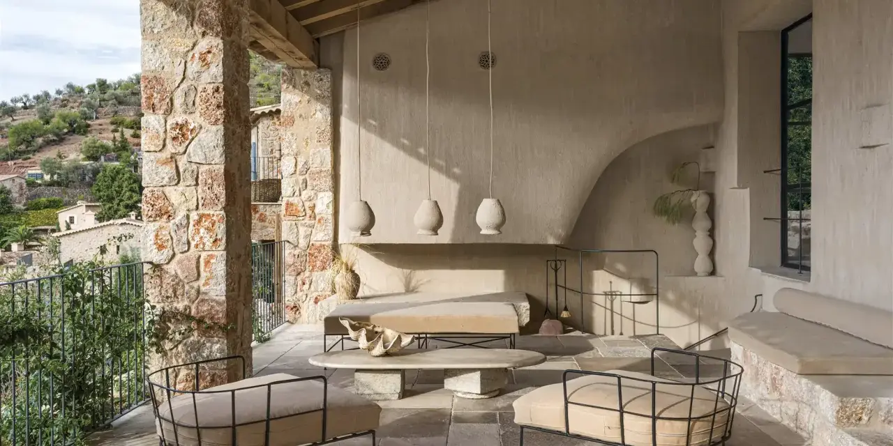 Acogedora terraza interior piso con sillones de metal, sofá y mesa de centro de piedra, bajo un techo de madera y rodeada de muros de piedra.