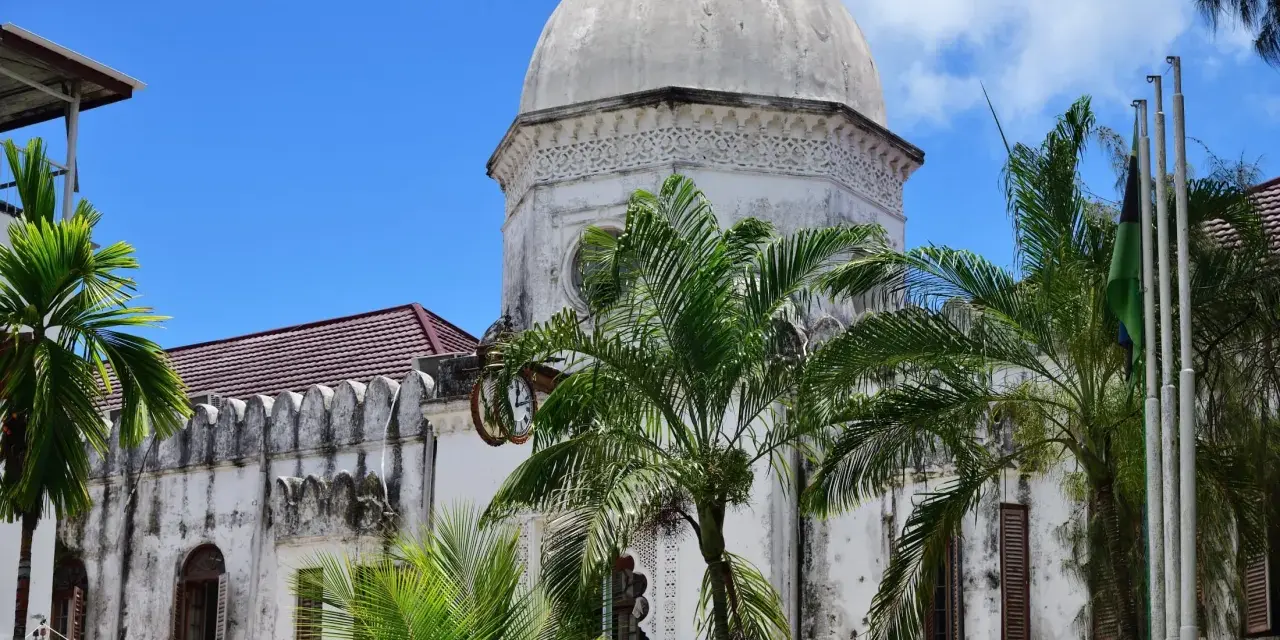 Biała, zabytkowa budowla z kopułą i palmami na tle błękitnego nieba. Klimatyczne zanzibar miasto.