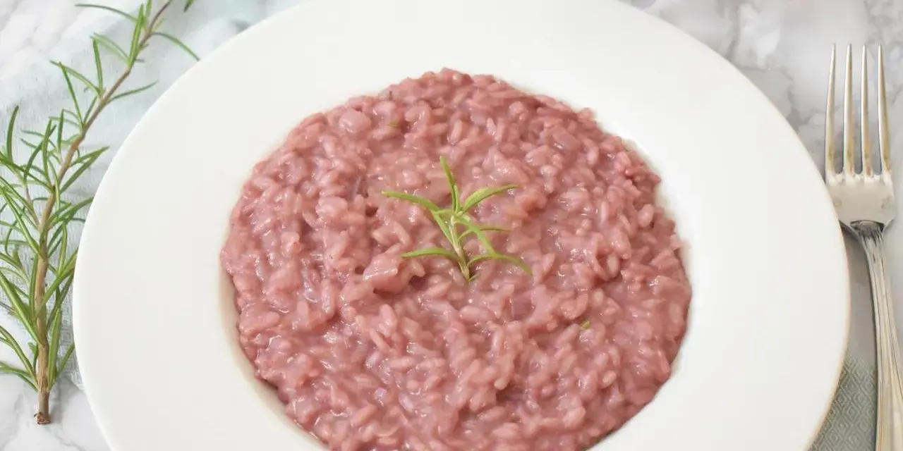 Różowe risotto z prawdziwkami i rozmarynem na białym talerzu, obok widelca i kieliszka wina.