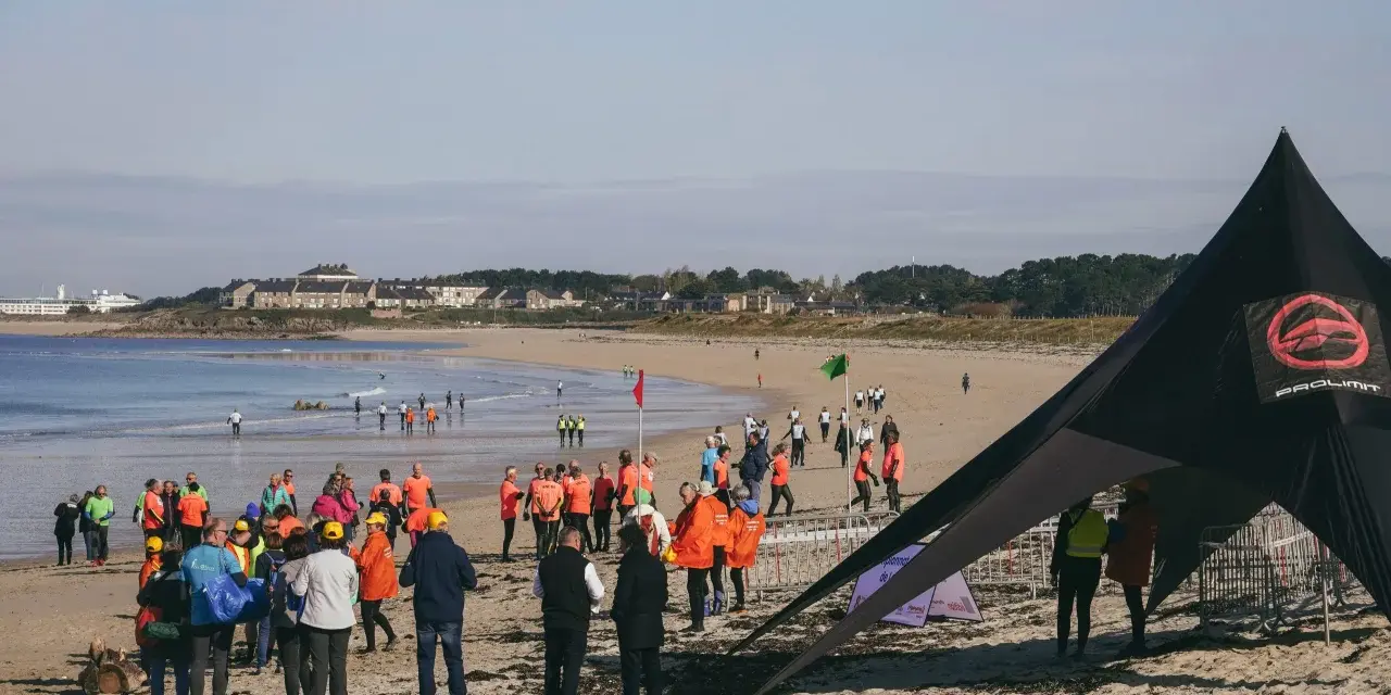 Rassemblés sur la plage pour une course de longe côte, des participants en tenues colorées attendent le départ sous un ciel clair.