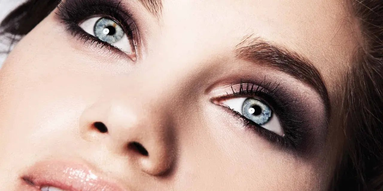 Occhi azzurri intensificati da un trucco smokey eyes audace, con labbra lucide e un accenno di sorriso.
