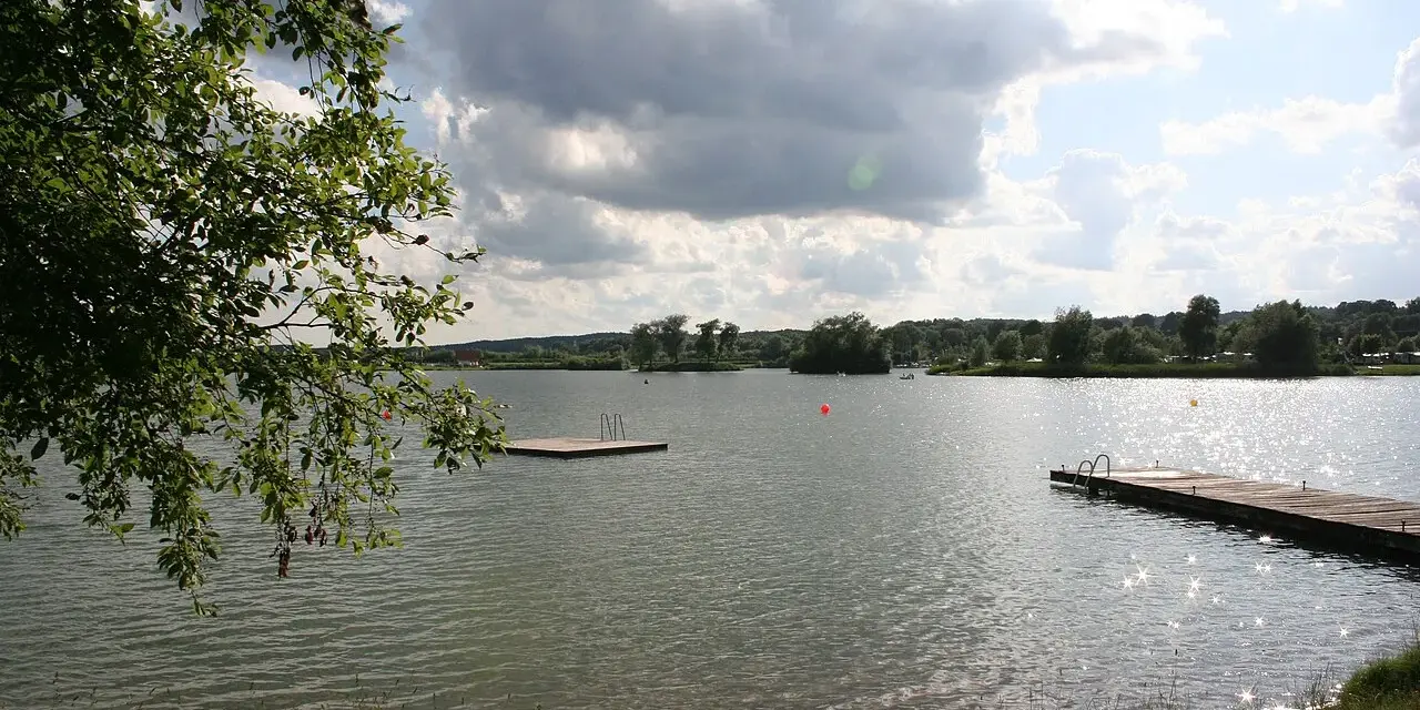 Ein sonniger Tag am Oberrieder Weiher Camping. Ein Steg mit Leiter lädt zum Baden ein, während im Hintergrund Bäume und eine idyllische Landschaft zu sehen sind.