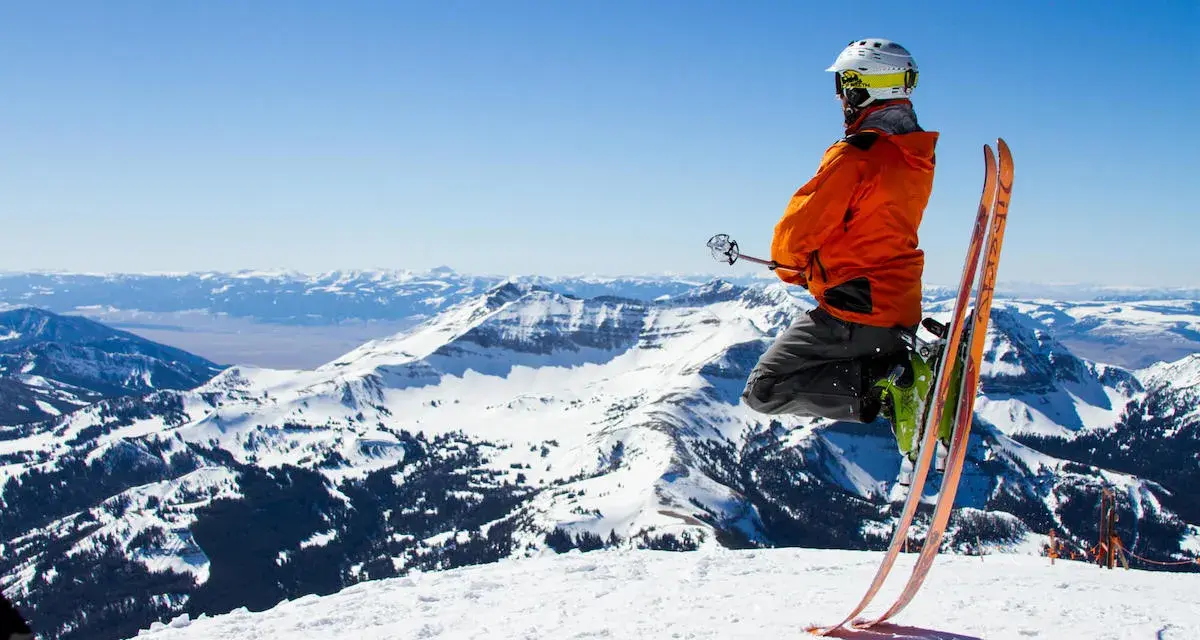 Un skieur en l'air, prêt à dévaler la pente. Il porte un casque et une veste orange vif, rappelant les règles pour skier en toute sécurité. Le paysage montagneux enneigé est spectaculaire.