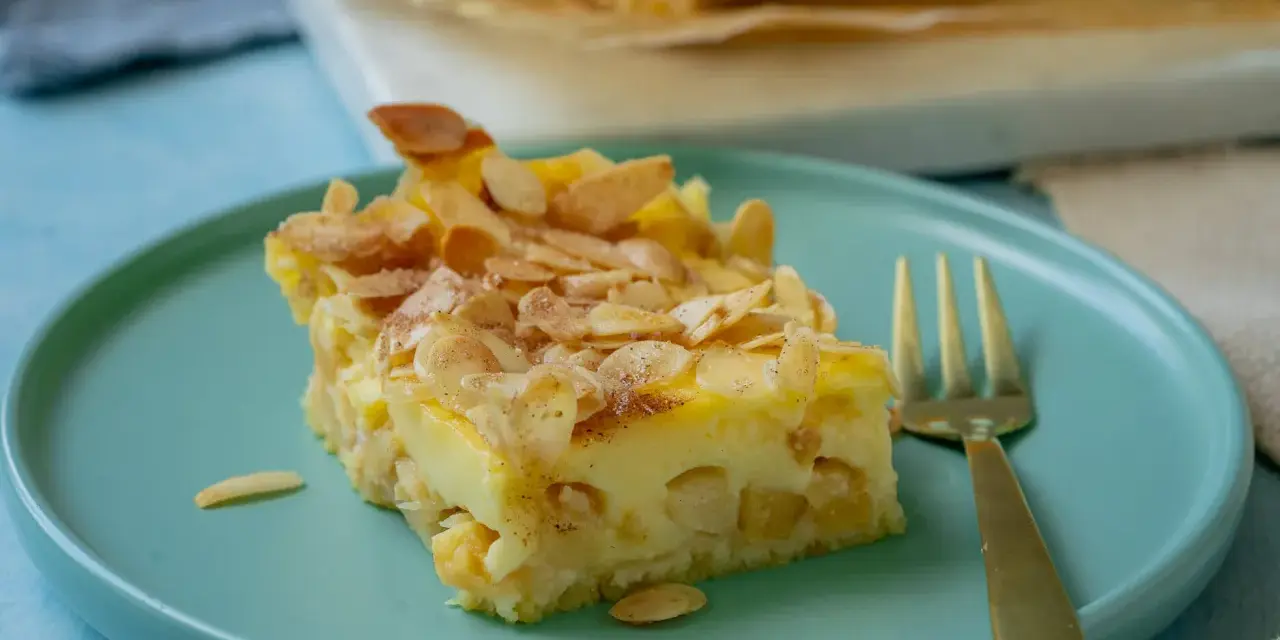 Ein Stück schneller Apfelkuchen mit Schmand und Pudding, bestreut mit Mandelblättchen und Zimt, auf einem türkisfarbenen Teller.