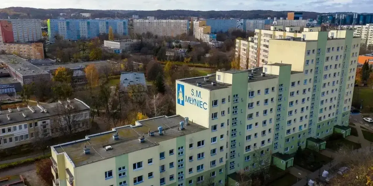 Duży blok mieszkalny S.M. Młyniec z charakterystycznym logo, otoczony innymi budynkami i drzewami jesienią.
