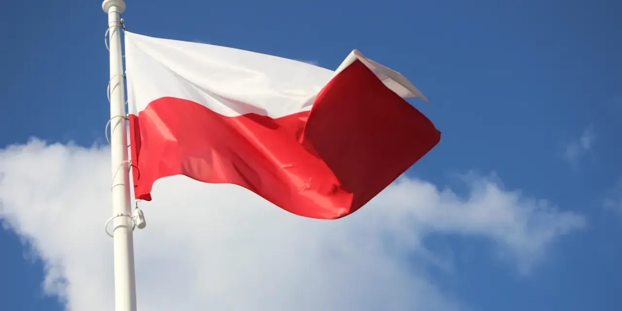 Biało-czerwona bandera polska powiewa na maszcie na tle błękitnego nieba z chmurami.