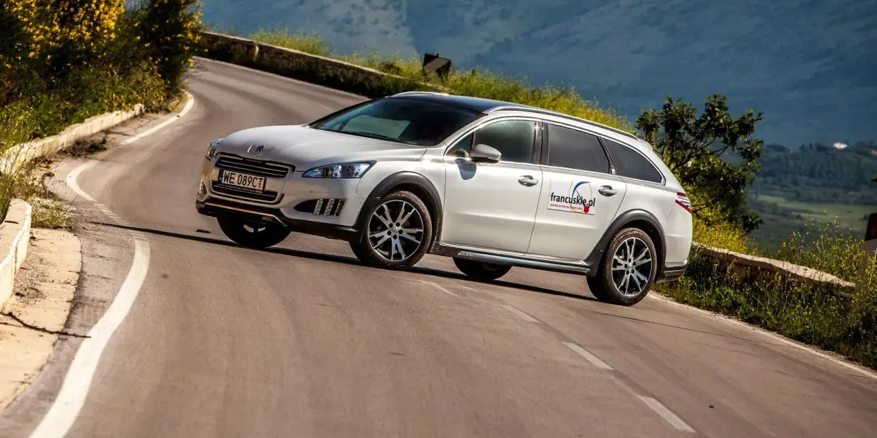 Białe kombi Peugeot 508 RXH na krętej drodze w górach.