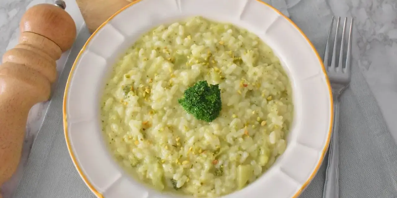 Kremowe risotto z brokułami, posypane pistacjami, z małą różyczką brokułu na wierzchu. Obok widelca i kawałków parmezanu.