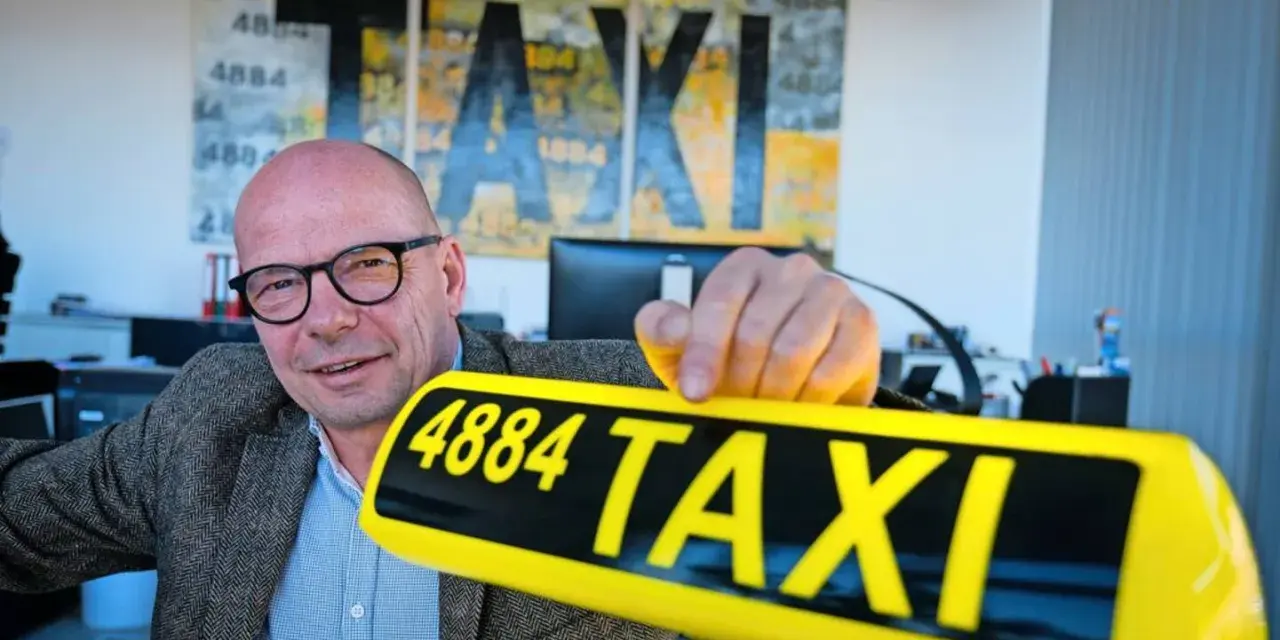 Mann mit Brille hält ein gelbes Taxi-Schild mit der Nummer 4884. Im Hintergrund Kunstwerke mit "TAXI" und der Zahl 1884.