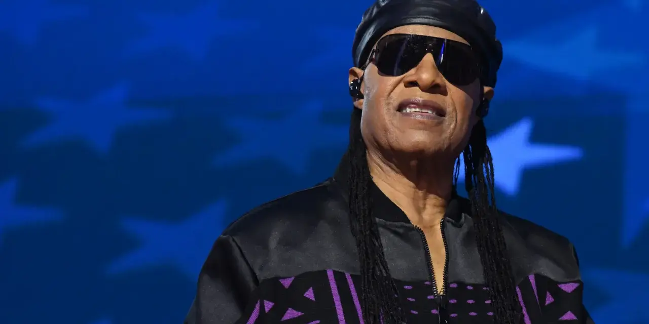 Stevie Wonder heute, mit Sonnenbrille und schwarzer Kappe, trägt eine Jacke mit lila Muster vor blauem Sternenhintergrund.