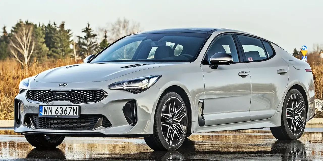 Elegancka Kia Stinger 2025 w kolorze srebrnym, zaparkowana na mokrej nawierzchni, z drzewami w tle.