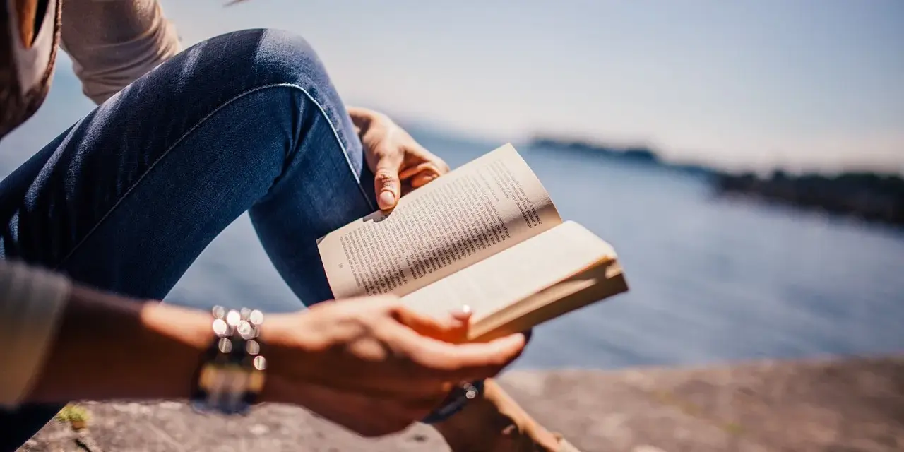 Persona sentada junto al agua, leyendo un libro en francés.
