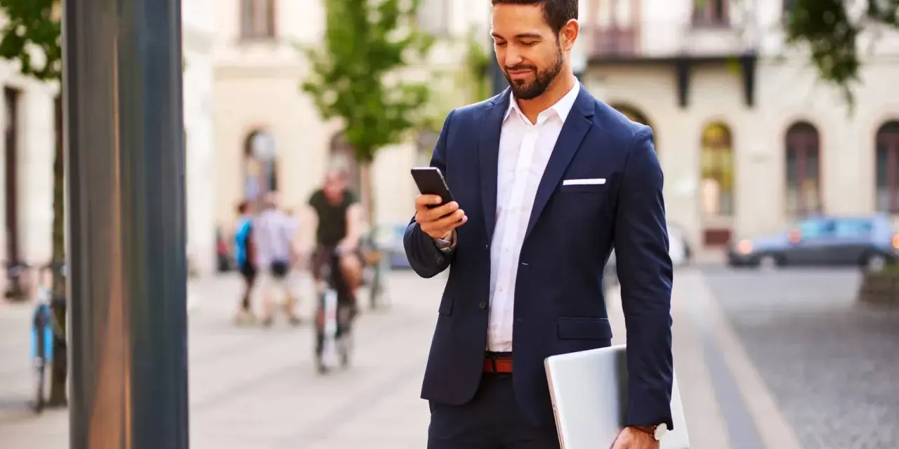 Mężczyzna w eleganckim stroju biznesowym męskim sprawdza telefon, trzymając laptopa.