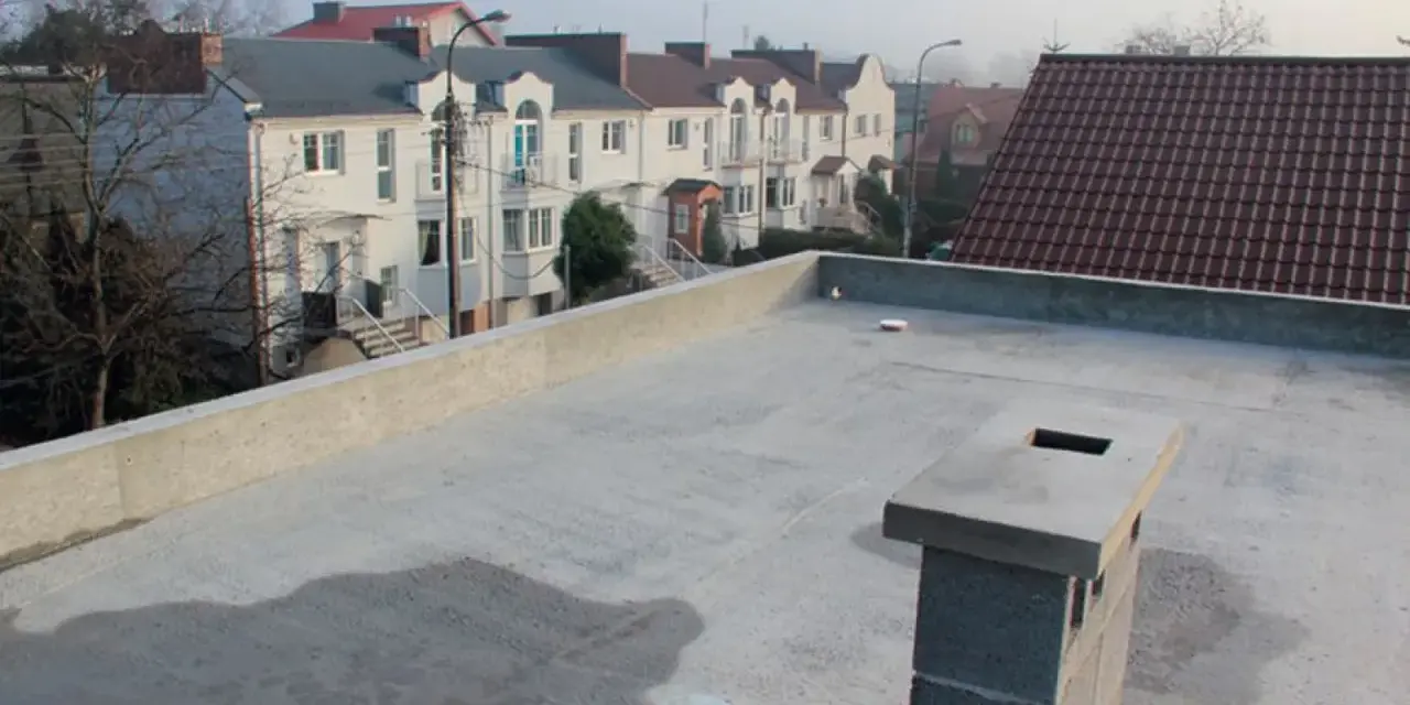 Nowy, betonowy dach płaski z kominem i widokiem na szereg domów z dachówką.