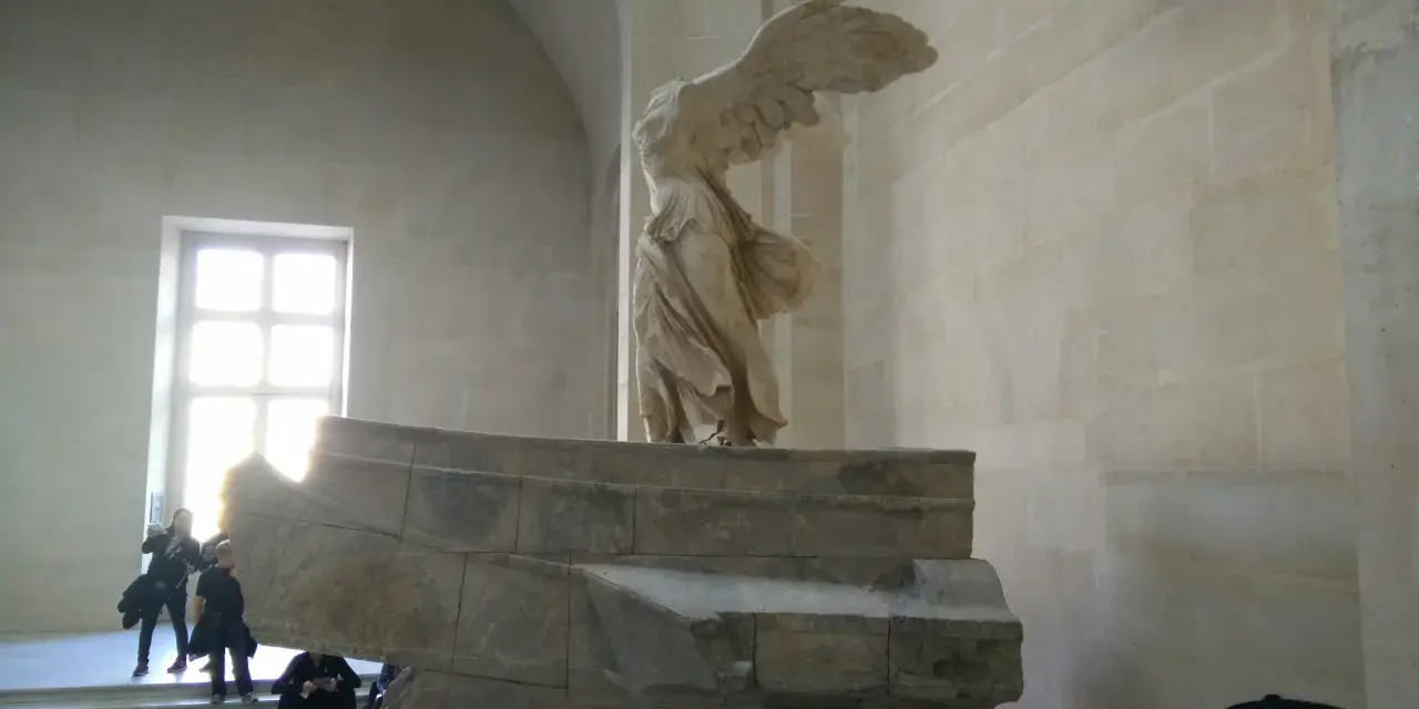 Skrzydlata Nike, symbol zwycięstwa, niczym mit, unosi się nad kamiennym postumentem.