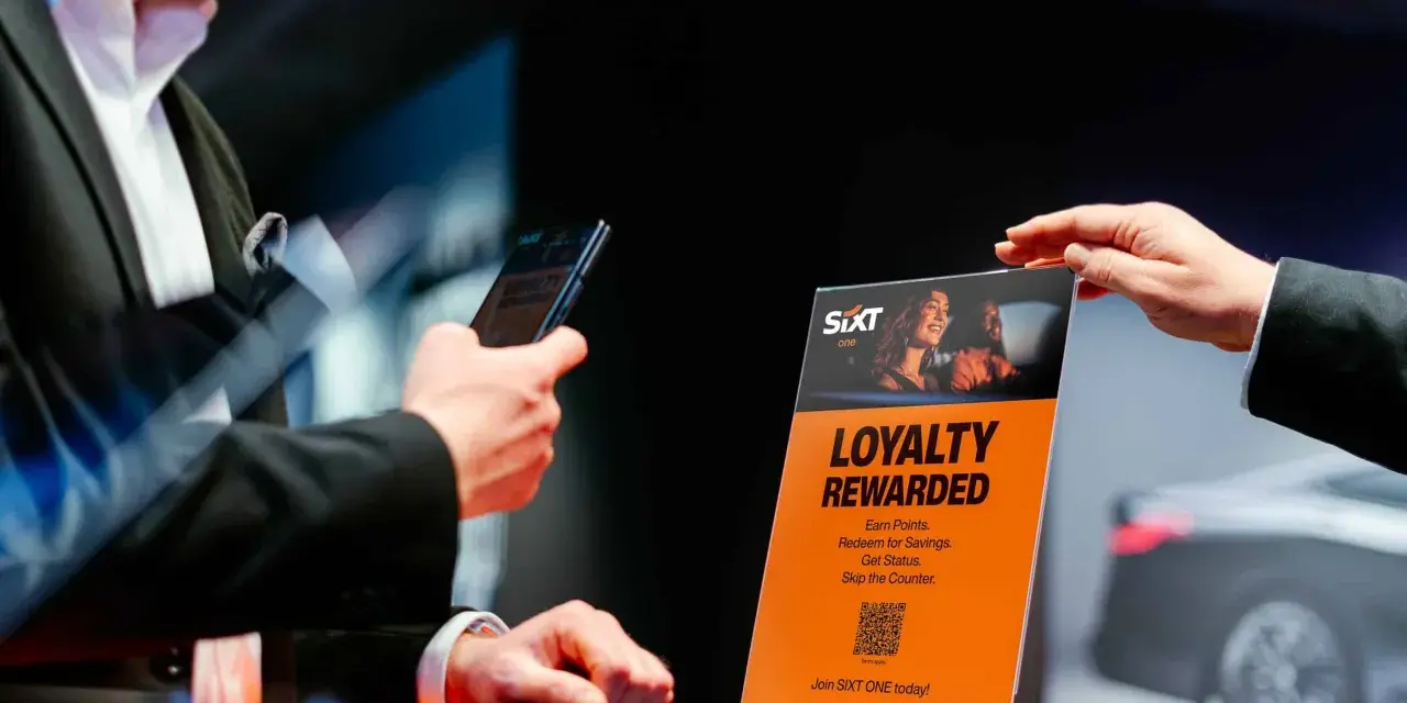 Ein Mann hält ein Smartphone, während eine Hand ein orangefarbenes Schild mit der Aufschrift "Loyalty Rewarded" und dem SIXT ONE Logo hochhält.