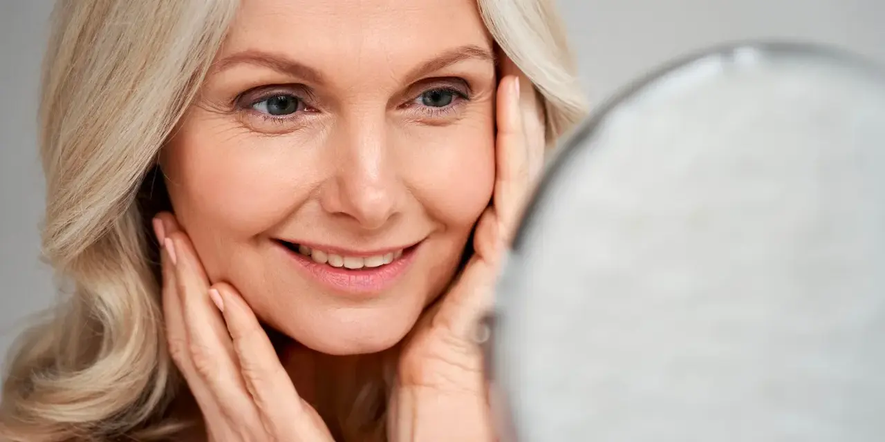Une femme blonde sourit en se regardant dans un miroir, ses mains caressant son visage. Le maquillage paupière tombante avant après est subtil.