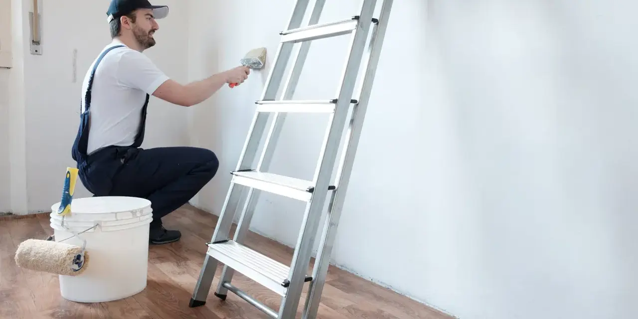 Hombre con overol azul y gorra, usando brocha para limpiar paredes. Escalera y cubo de pintura a su lado.