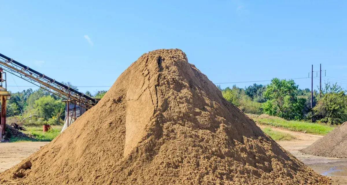 Ogromna hałda piasku na placu budowy, zastanawiasz się, ile kosztuje tona piasku? W tle taśmociąg i drzewa.