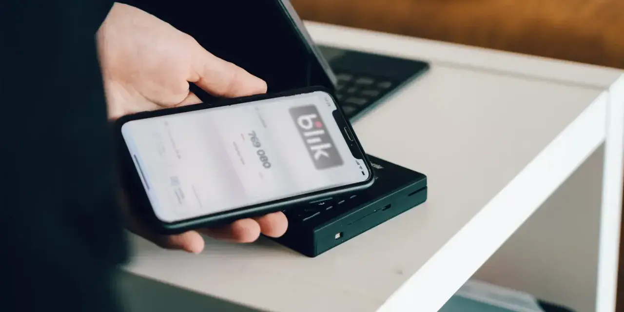 Dłoń trzyma smartfon z aplikacją Blik i kodem transakcji nad terminalem płatniczym.
