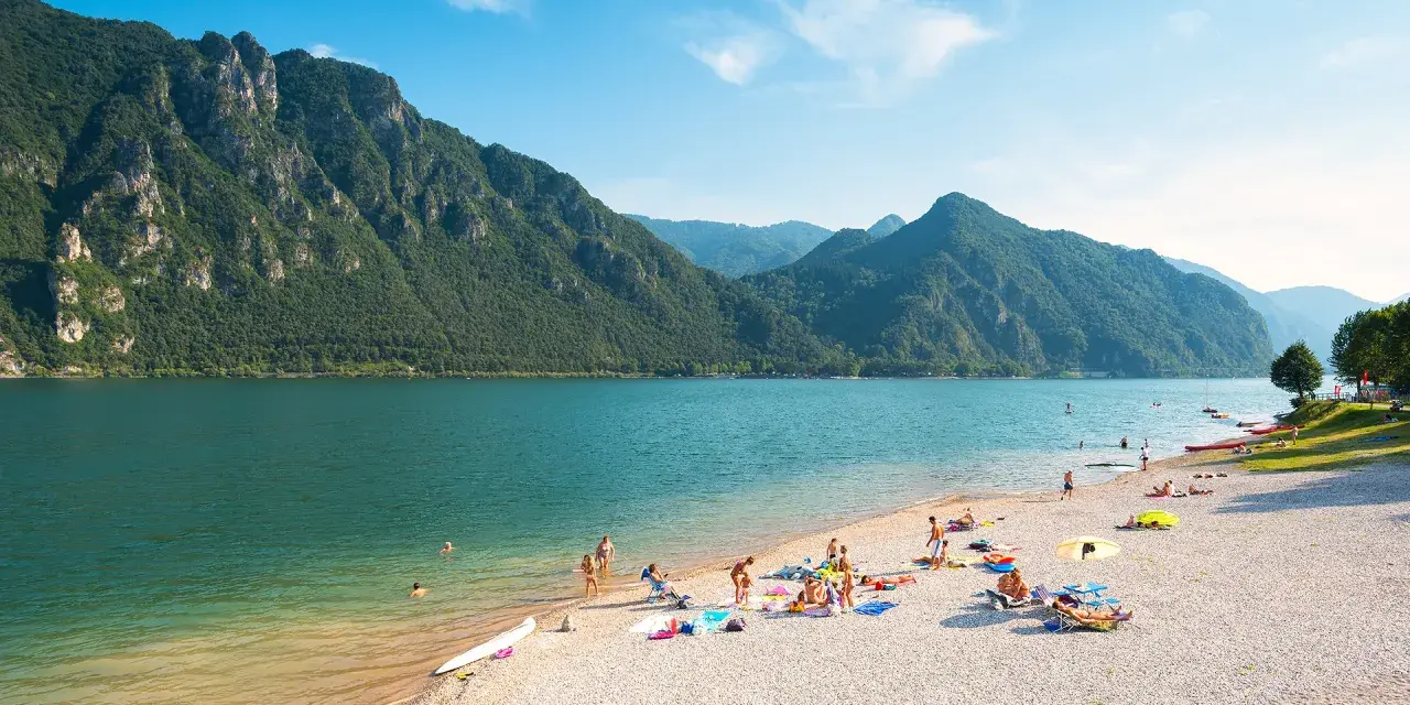 Spiaggia affollata sui laghi in Piemonte, ideale per una gita in moto. Montagne verdi e acqua azzurra.