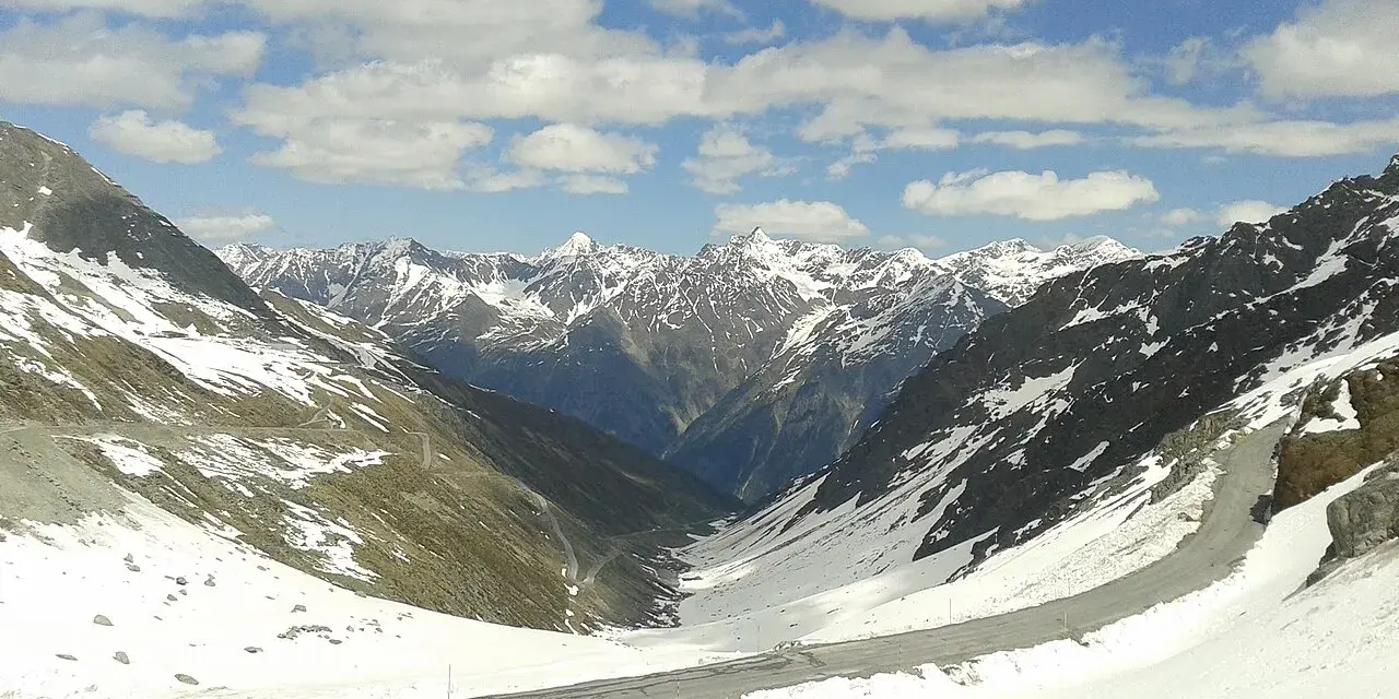 Strada tortuosa sulla Ötztaler Gletscherstraße, tra montagne innevate e cielo azzurro con nuvole bianche.