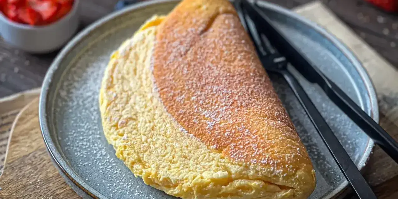 Puszysty omlet, lekko przyprószony cukrem pudrem, czeka na talerzu obok czarnych sztućców. W tle miseczka z truskawkami.