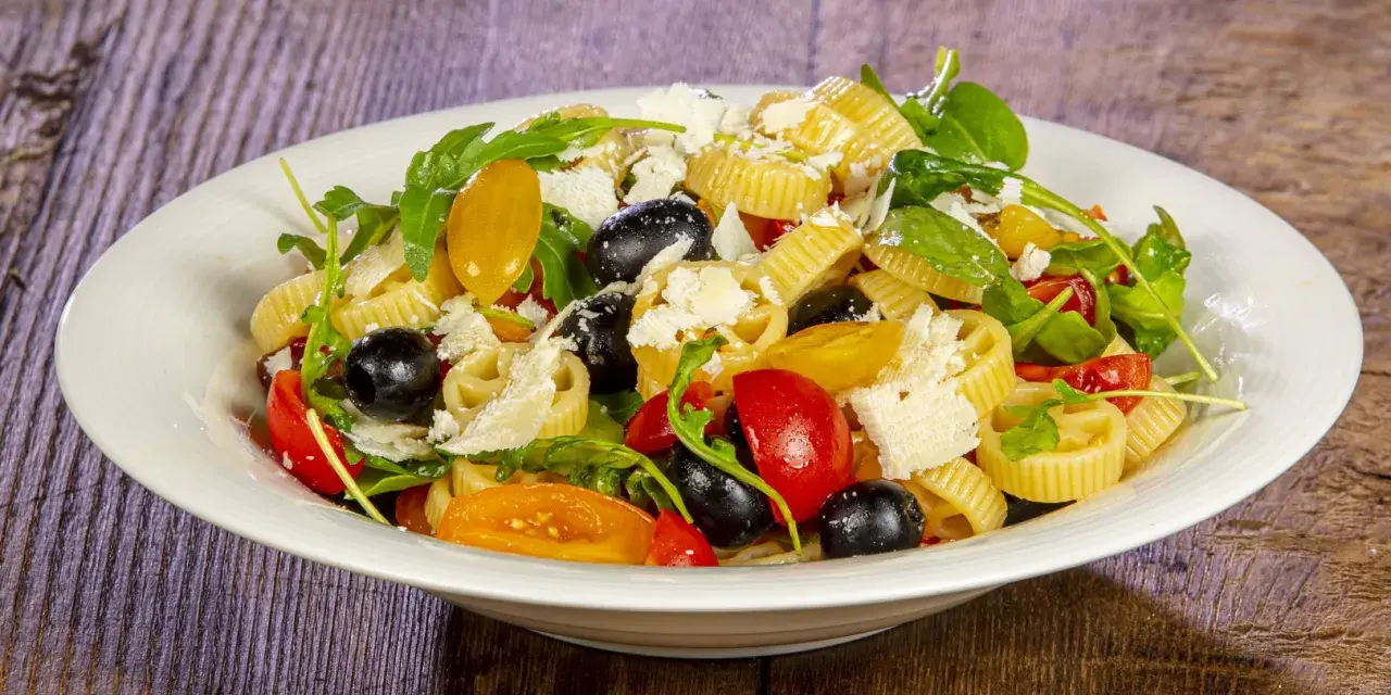 Un piatto di pasta alla crudaiola con pomodorini, olive nere, rucola e scaglie di parmigiano. Fresca e colorata.