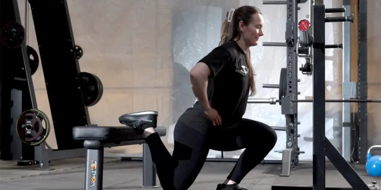 Kvinna utför en split squat i ett gym. Hennes ena fot vilar på en bänk bakom henne.