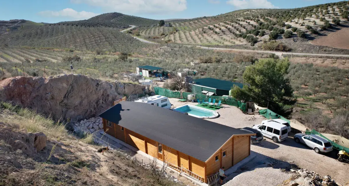 Casa de madera con piscina y caravana en terreno rústico, rodeada de olivos y colinas. Ideal para quienes buscan un refugio natural.