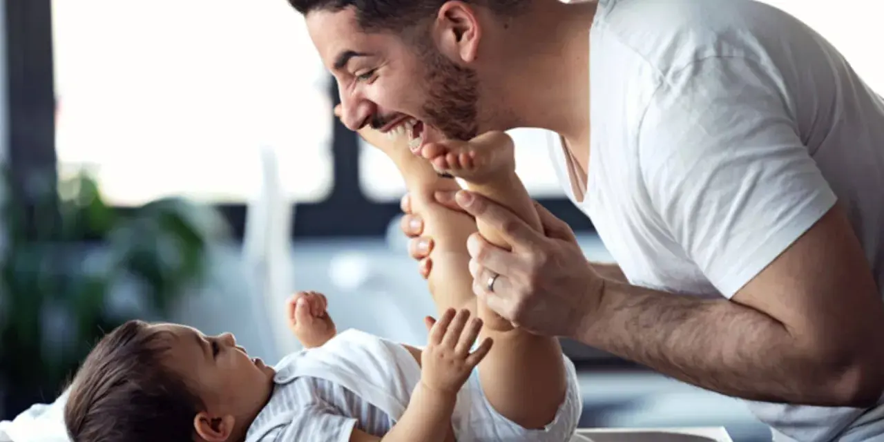 Ein lachender Vater mit Kind spielt liebevoll mit den Füßen seines Babys.