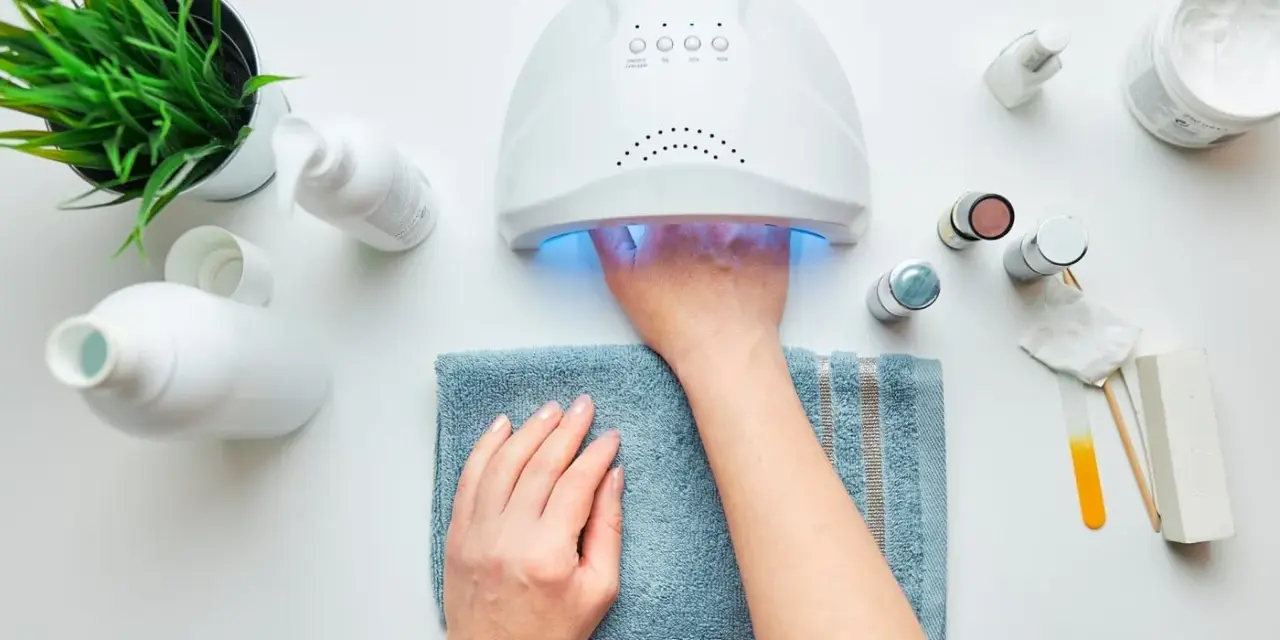 Paznokcie hybrydowe krok po kroku: dłoń w lampie UV, obok kosmetyki i akcesoria do manicure.