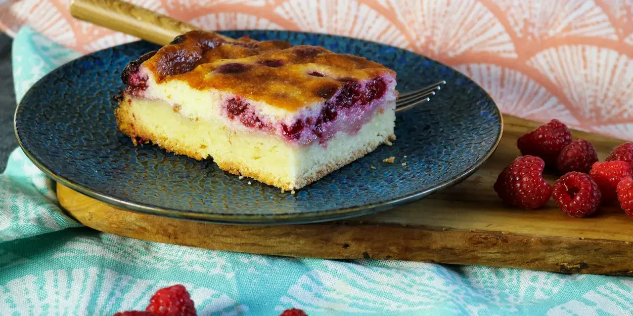 Ein Stück Himbeer Schmand Kuchen auf einem blauen Teller, daneben frische Himbeeren.